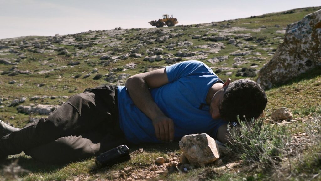 No Other Land film still, NYFF 2024. Man lies on rocky hillside, bulldozer in distance. Documentary film.