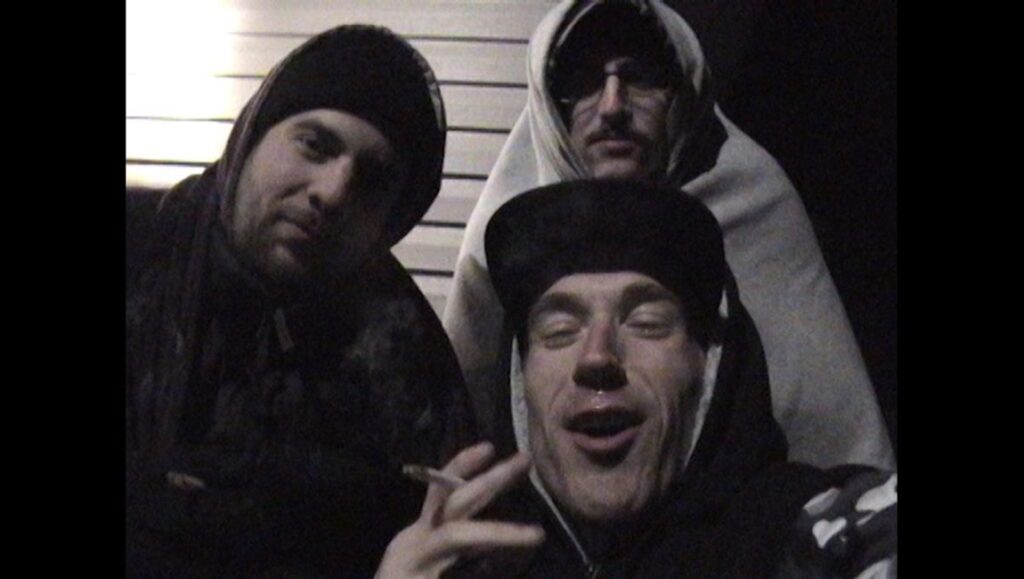Rap World film still for New/Next Film Fest 2024. Three men, one smoking, in a grainy, dark scene.