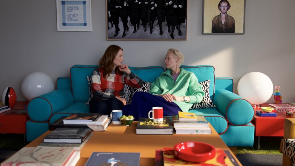 The Room Next Door NYFF 2024 film still: Julianne Moore and Tilda Swinton on a teal couch in a colorful room.