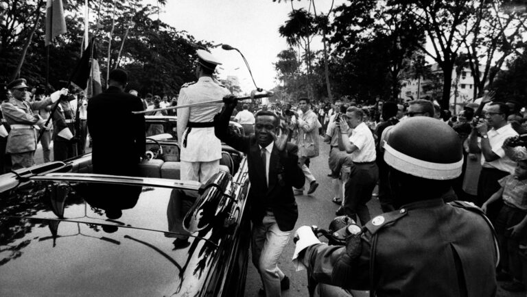 Black and white image from Soundtrack to a Coup d'État featuring a parade with crowds and officials.