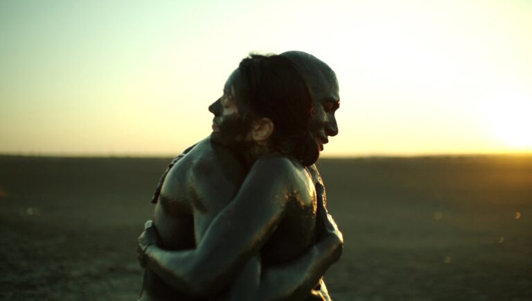 The Black Sea movie still: Derrick B. Harden and Irmena Chichikova embrace, covered in mud, against a sunset backdrop.
