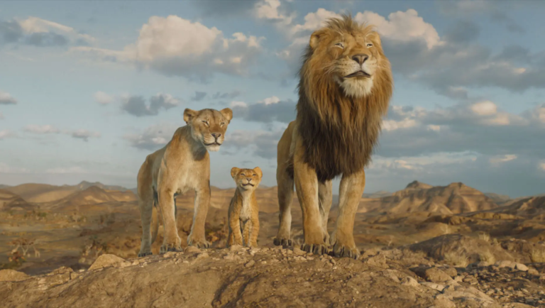 Mufasa: The Lion King movie still. A lion, lioness, and cub stand on a rocky outcrop against a desert backdrop.