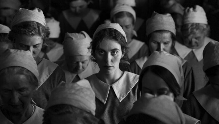Black and white still from The Girl with the Needle movie. Woman stares intensely amidst a crowd in uniform.