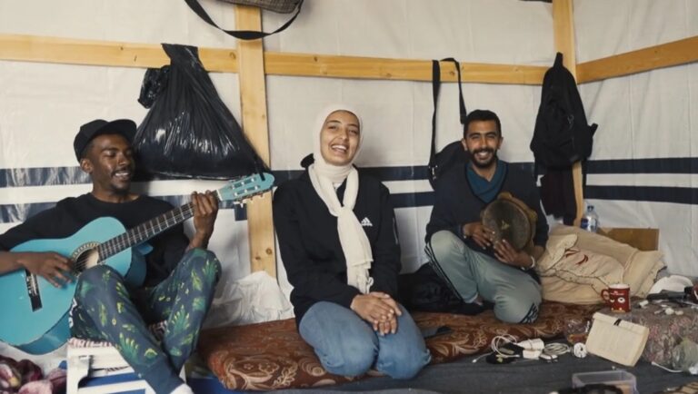 From Ground Zero film scene. Musicians play guitar and percussion, smiling. Rashid Masharawi film review.