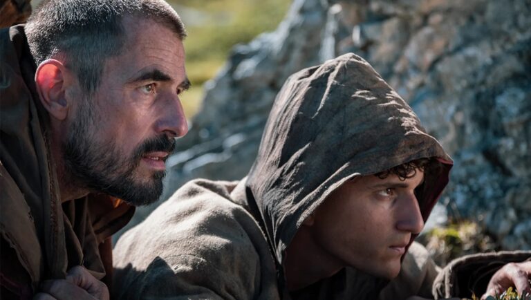 Film scene with two men. Older man with beard, younger hooded character. Drama, period piece.