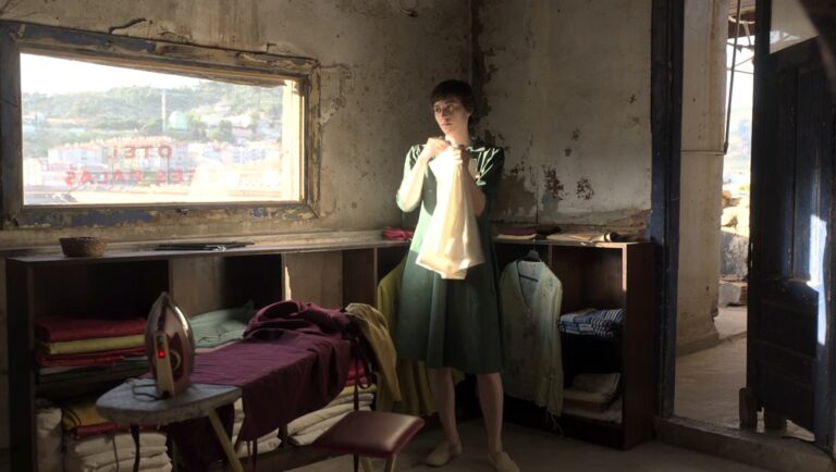 Film still of woman in vintage dress holding clothing in aged room. Iron and linens visible. And the Rest Will Follow movie.