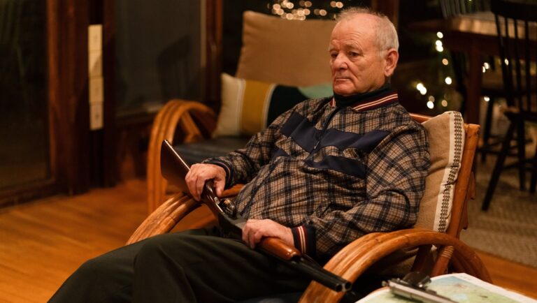Bill Murray in Riff Raff. Actor seated, holding a rifle, wearing a plaid shirt. Movie scene.
