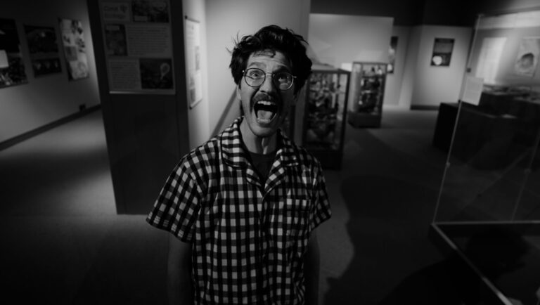 Cory Santilli in In The Mouth. Black and white film still of a man with glasses screaming in a museum.