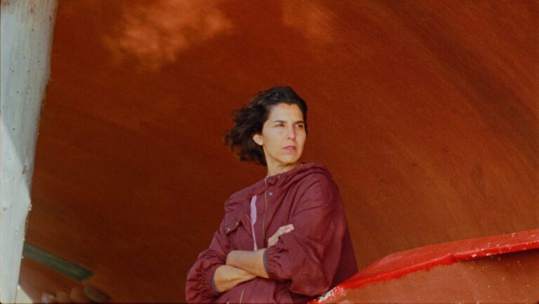 Ariel IFFR25 film still. Woman with crossed arms, wearing a maroon jacket. Red and orange background.