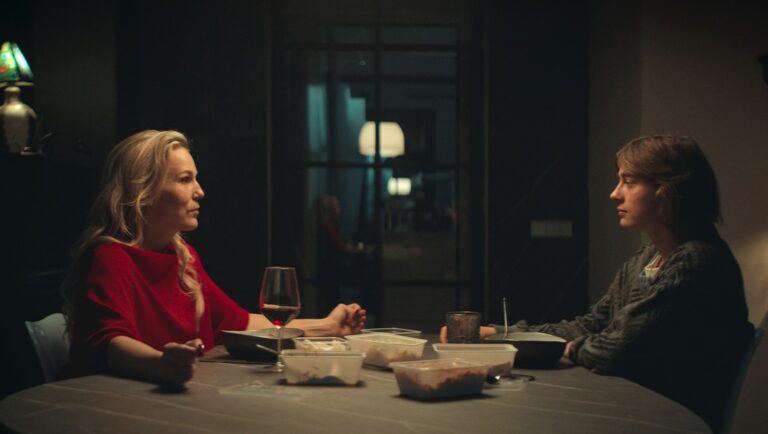Two people at a table. Woman in red, person in grey. Food containers, wine glass, and a mug. Indoor scene.