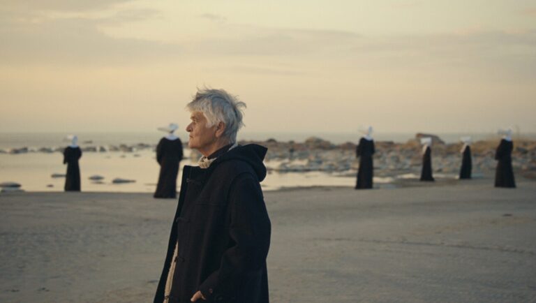 Orenda film still. Woman in coat on beach with figures in nun habits. IFFR 2025.