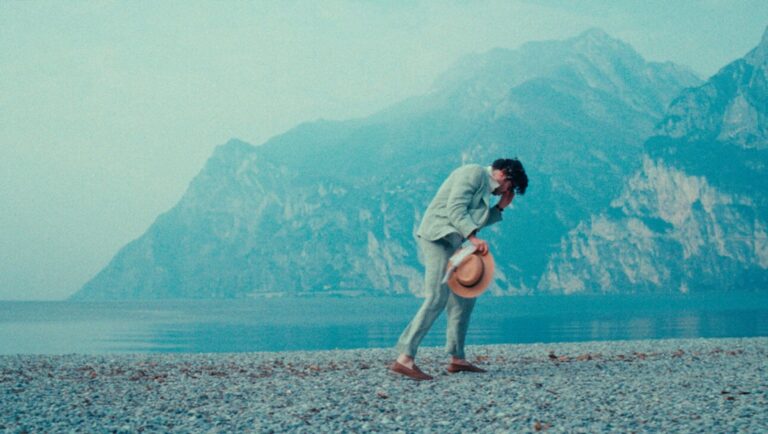 IFFR 2025 — Dispatch 3: Pavements, The Last Dance, The Things You Kill Man on pebble beach with mountains. Man in suit holding hat. Lake Garda scenery. Travel.