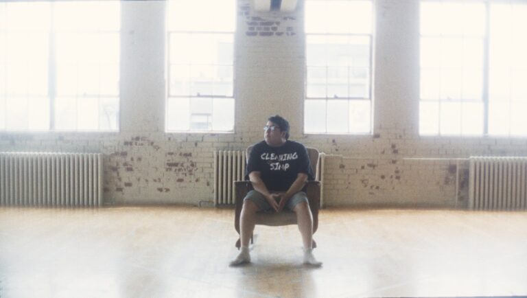 Man in Cleaning SImp T-shirt sits in chair. Bright room with large windows, brick walls, and radiator. Film still.