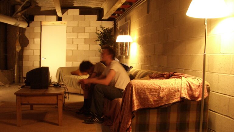 Two men in a basement apartment. Retro TV, cinder block walls, and cozy, dim lighting. Wheelhouse Creative.