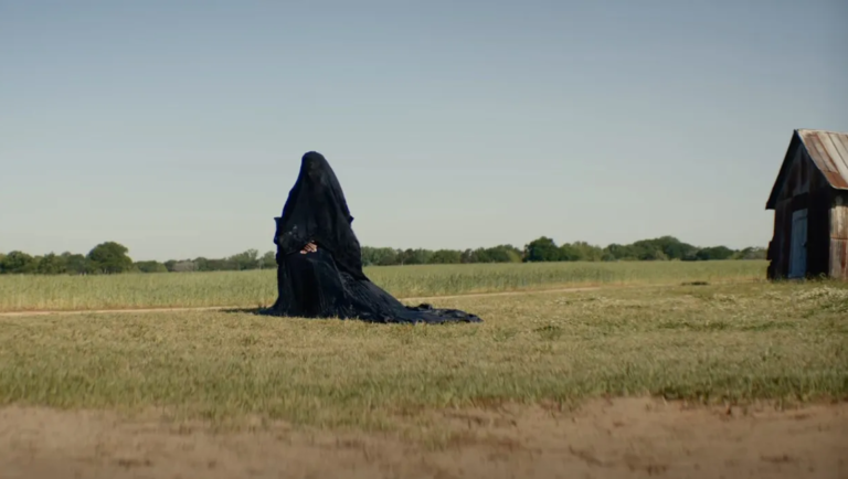 The Woman in the Yard movie scene. Figure in black fabric stands in a field near a shed. Jaume Collet-Serra film review.