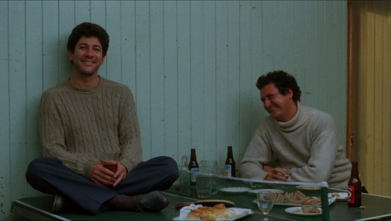 Peter Riegert in Local Hero film still. Two men sit at a table with beer and food. Criterion Collection.