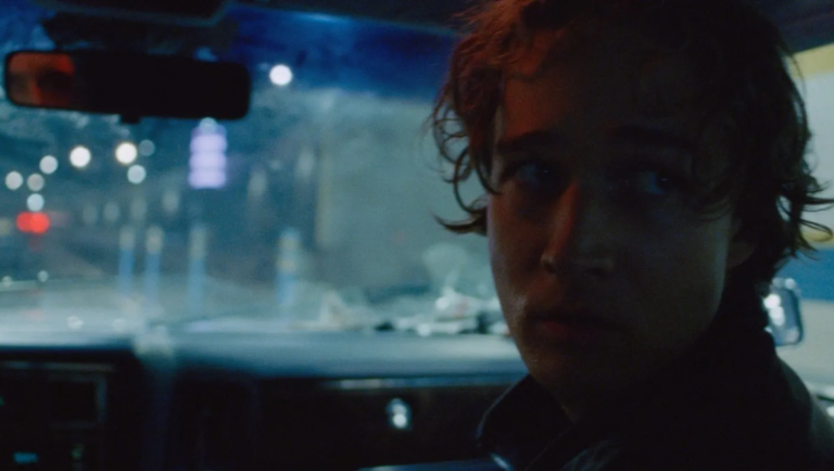 Pet Shop Days movie scene: A young man in a car, looking concerned, with city lights blurred in the background.