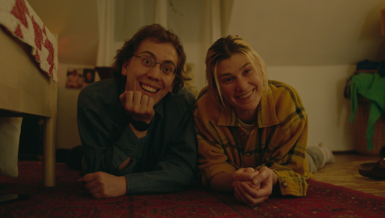 Two smiling people lie on a rug. The man wears glasses and a blue shirt. The woman wears a yellow flannel.