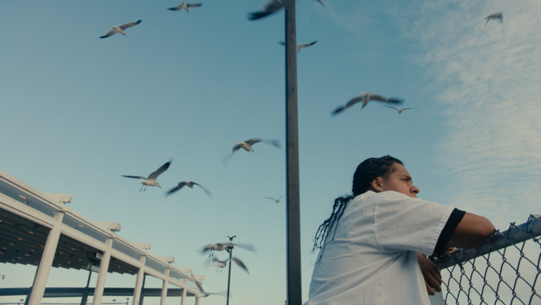 Mad Bills to Pay film still. Person by fence under seagulls. New Directors/New Films 2025.