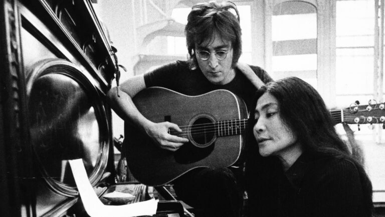 John Lennon and Yoko Ono in black and white. Lennon plays guitar, Ono stands beside him.