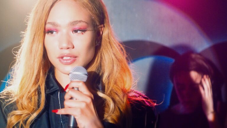 Singer Louiza Aura performing with a microphone. Stage lights, pink eyeshadow, and red nails.