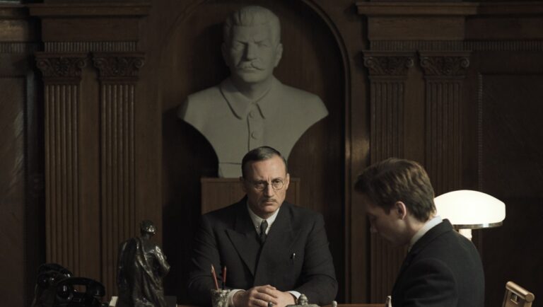 Two Prosecutors film still from Cannes Film Festival 2025. Man in suit seated below bust.
