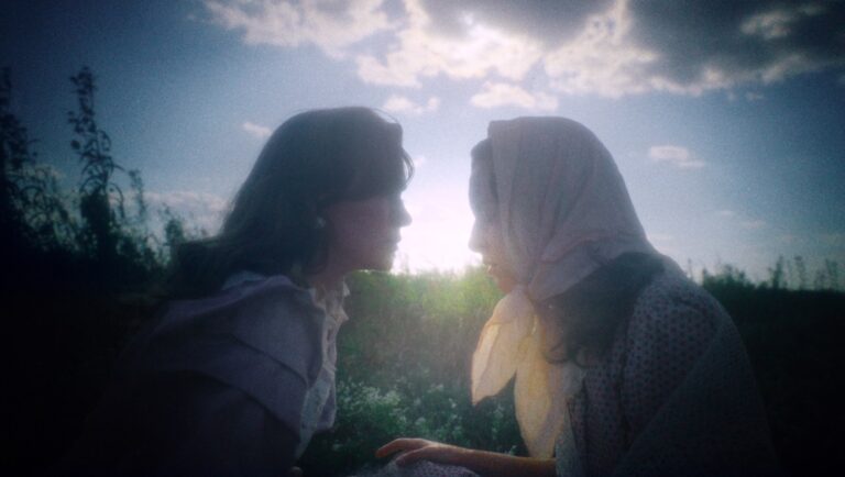 Two women face each other in An Evening Song movie still. Headscarf, sunlight, and outdoor scene.