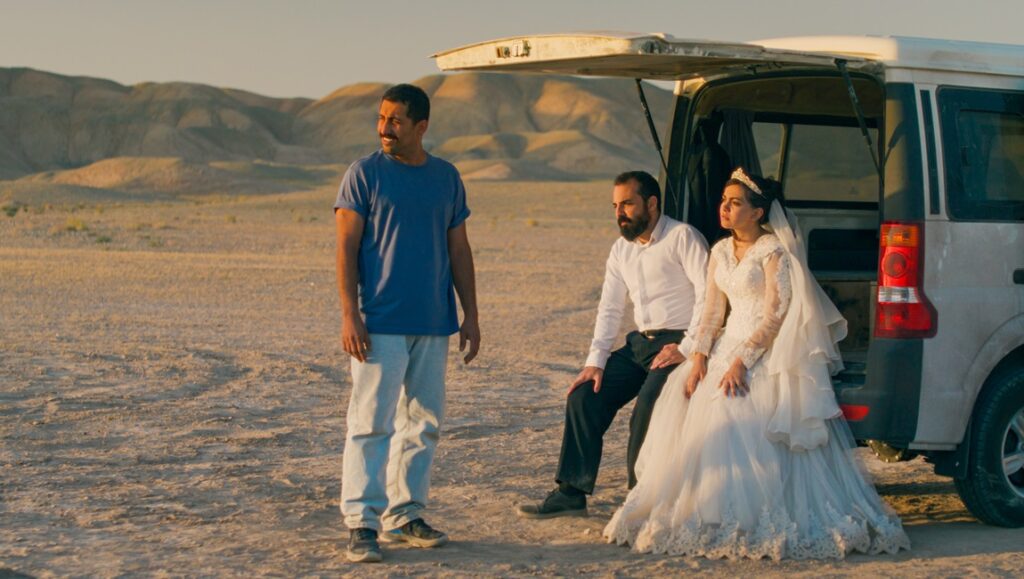 Wedding in the desert. Bride in white dress, groom, and man by car. Just an Accident film scene.