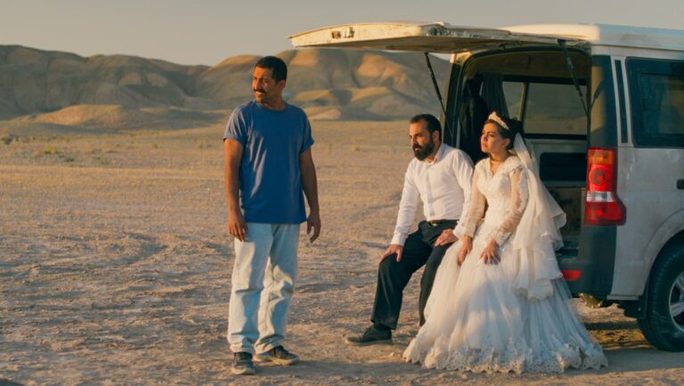Wedding in the desert. Bride in white dress, groom, and man by car. Just an Accident film scene.