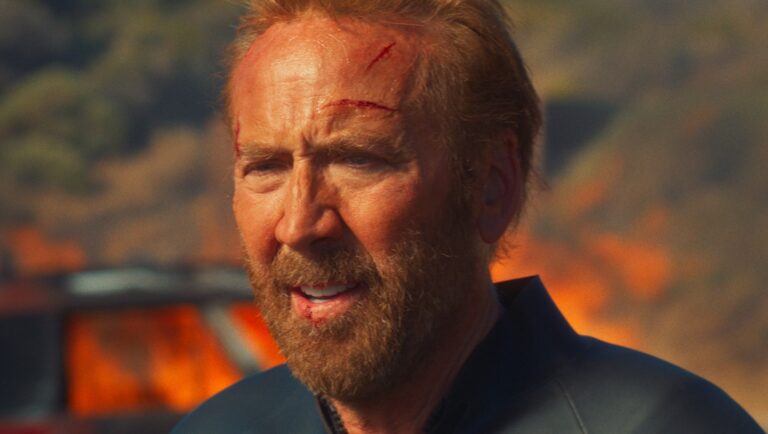 Nicolas Cage in The Surfer movie. Close-up of Cage with a cut on his forehead, beard, and burning wreckage in the background.