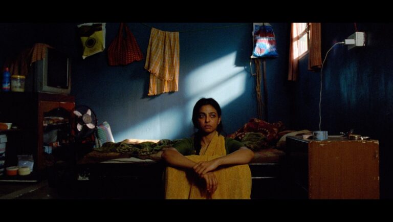 Sister Midnight movie still: Woman sits in a dimly lit room with blue walls. Bags hang from the ceiling. Film, drama, indie cinema.