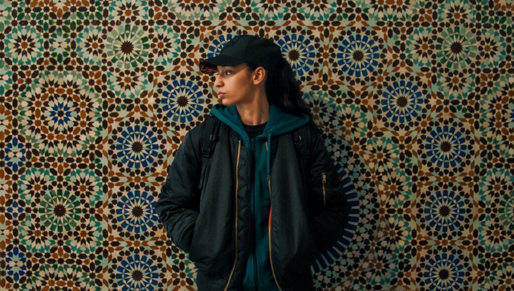 Hafsia Herzi in The Little Sister film. Woman in cap and jacket against a patterned wall. Cannes '25 review.