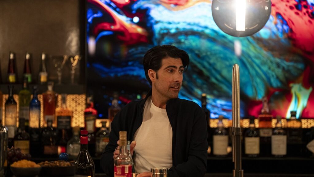 A Fundamental Absence in the Techno-Paradise Project — Jesse Armstrong’s Mountainhead Bartender at work. Man stands behind a bar with liquor bottles and a colorful backdrop.