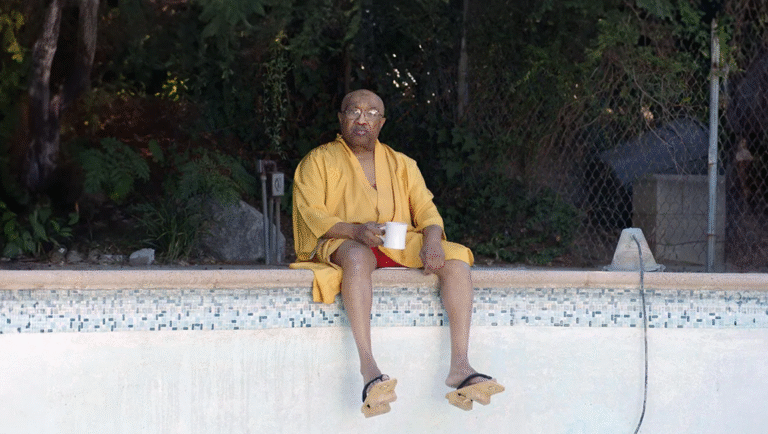 Swamp Dogg relaxing by the pool. Man in robe and sandals, holding a mug. Poolside relaxation.