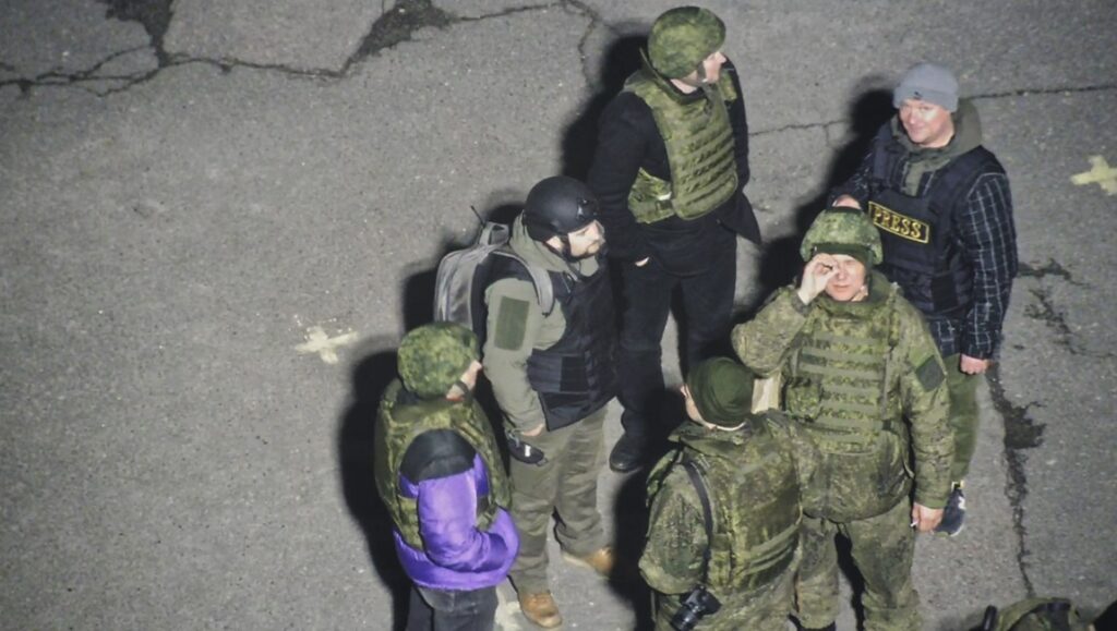 A group of six people, some in military uniforms and body armor, stand together outdoors on a paved surface, conversing as if preparing for a film interview. Two wear press vests and helmets. The scene appears to be at night or in low light.