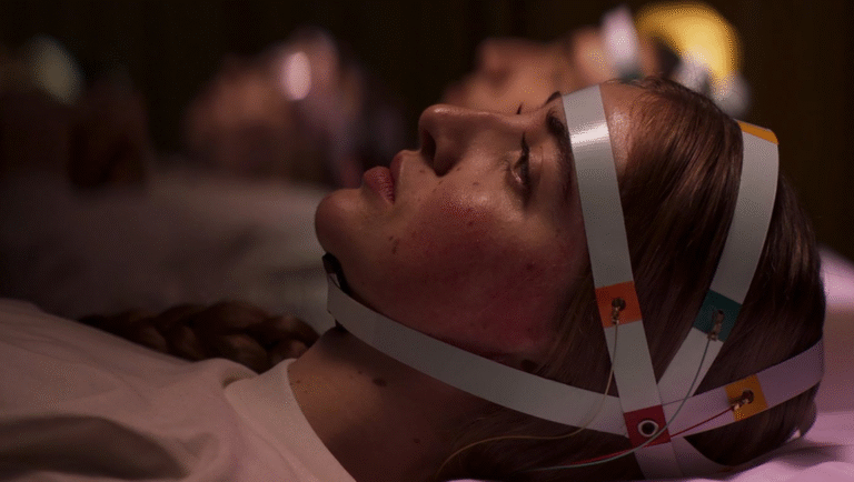 A young woman lies on her back wearing a white head strap with colored sensors, appearing to undergo a medical or scientific procedure. Other people wearing similar headgear are blurred in the background.