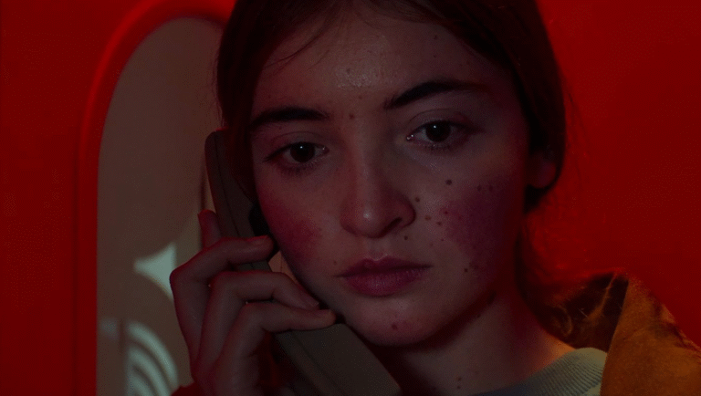 A young person with flushed cheeks and freckles holds a beige phone to their ear, looking serious under red lighting—capturing an intimate moment perfect for film festival coverage. A light, curved shape is visible in the background.