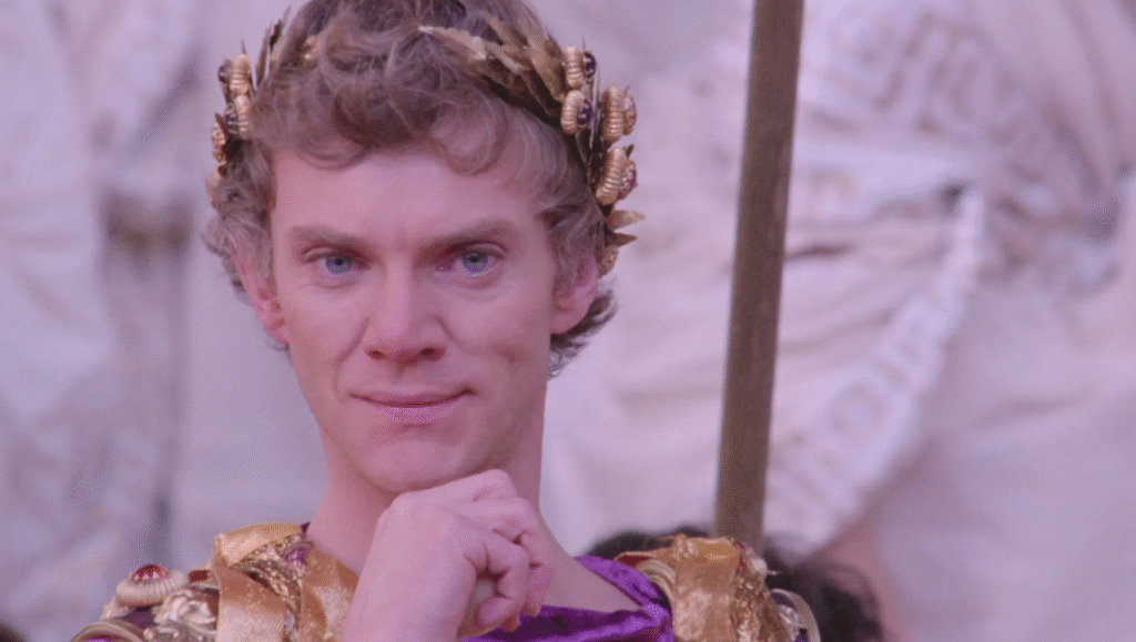Malcolm McDowell as Caligula staring straight ahead with his hand on his chin.