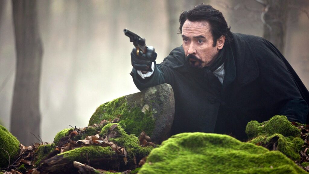 A man in a black coat pointing a pistol while leaning on a green hill.