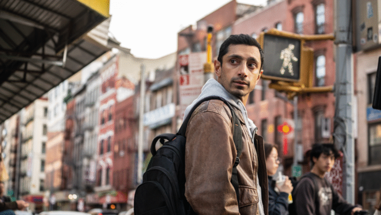 A man wearing a brown jacket and black backpack stands on a city street corner, looking over his shoulder. Urban buildings and a lit walk sign are visible in the background, along with several pedestrians.