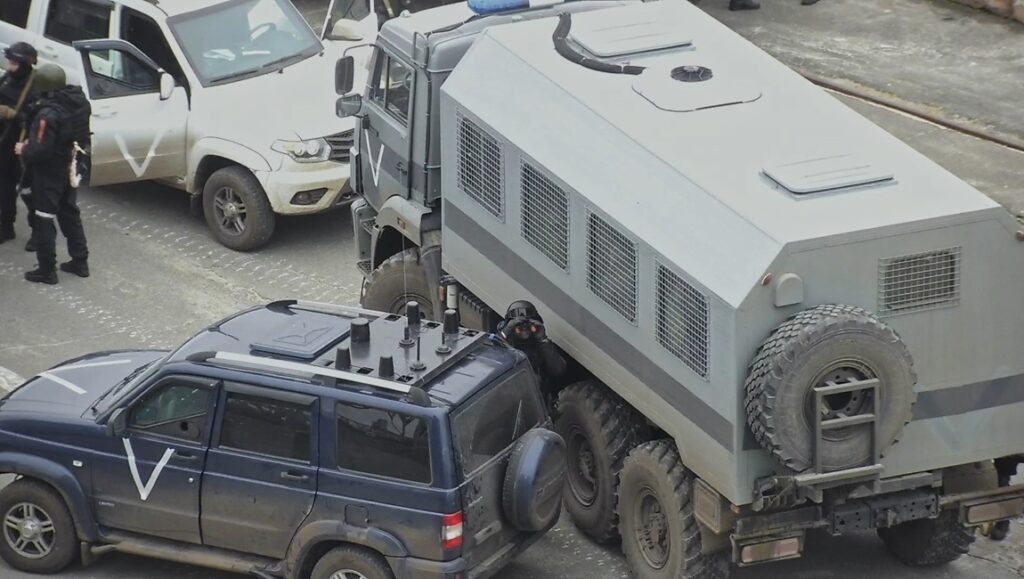 Several military vehicles, including an armored truck and two SUVs marked with white V symbols, are parked on a street where a film interview appears to be underway. Armed individuals dressed in tactical gear stand nearby.
