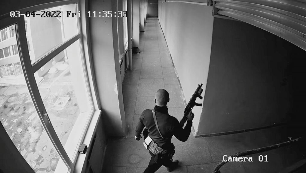 Surveillance camera footage, similar to behind-the-scenes shots from a film interview, shows a person in dark clothing carrying a rifle walking down a hallway with windows on the left. The timestamp reads 03-04-2022 Fri 11:35:33 and Camera 01 is displayed in the corner.