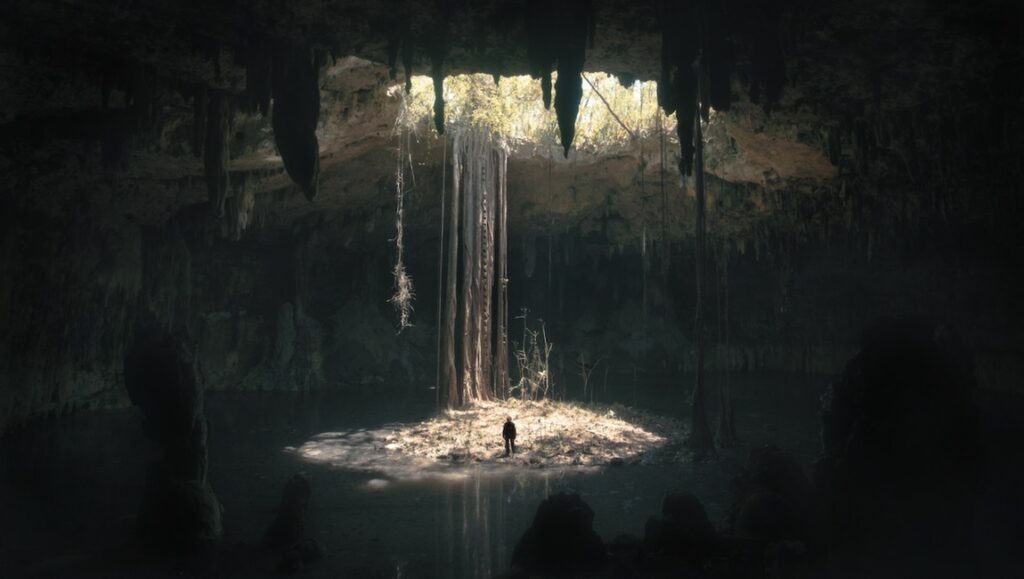 An underground cave with stalagmites and water, and a man standing in a ray of light coming into the cave.