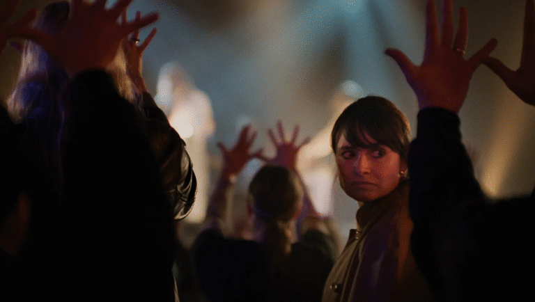 A person in a crowd looks over their shoulder while others around them raise their hands with spread fingers toward a stage lit by dramatic, colorful lighting.
