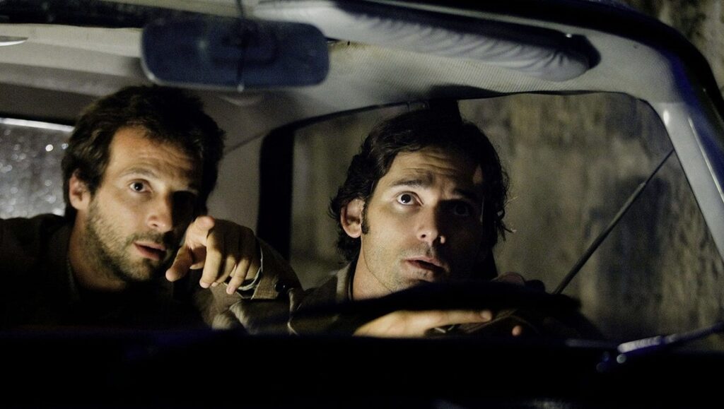Two men sit in a car at night; the man in the passenger seat points ahead with concern, while the driver looks intently forward, gripping the steering wheel.