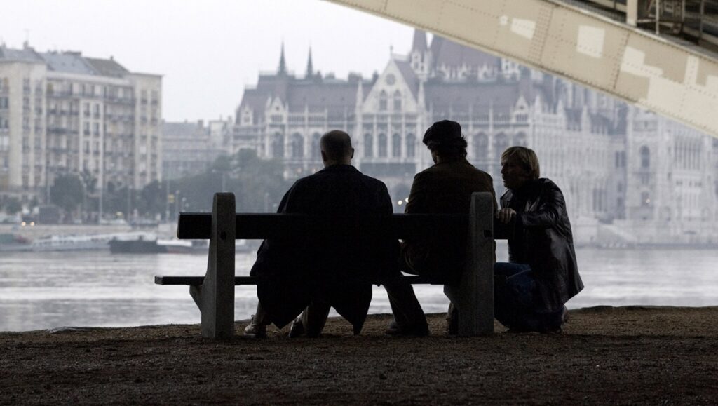 Three people sit on a bench by a riverbank, facing away, with historic buildings and a bridge in the background on an overcast day.