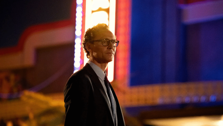 A man in a suit and glasses stands outside at night, illuminated by colorful neon lights and a glowing sign, creating a dramatic, cinematic atmosphere.