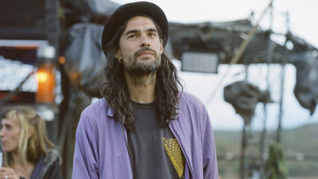 Óliver Laxe portrait. The director of 'Sirât' wears a hat and purple jacket. Indie film.