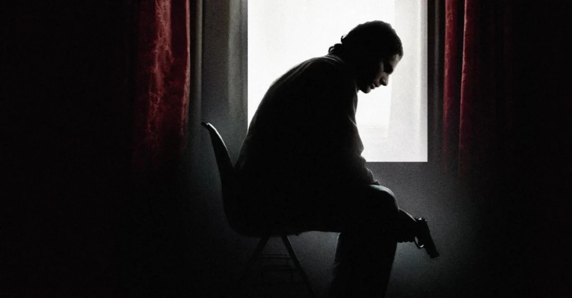 Breaking Bread: Steven Spielberg’s Munich A person sits on a chair in front of a bright window, silhouetted in shadow, holding a handgun with their head bowed.
