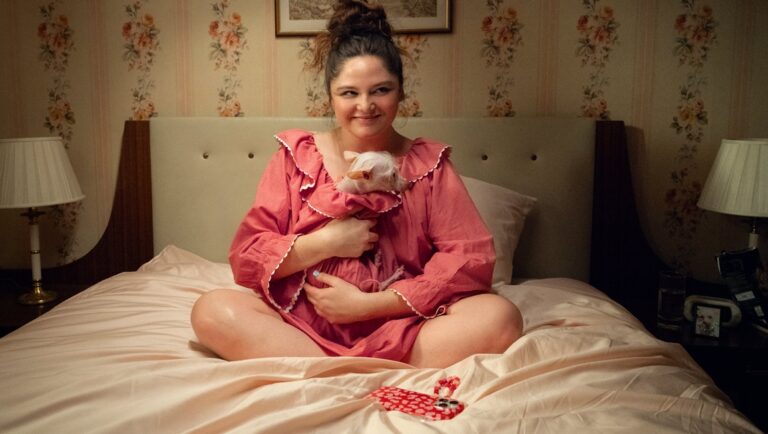 Ana Blumenkron sits cross-legged on a bed, cradling a piglet in her arms. Pink nightgown, floral wallpaper.